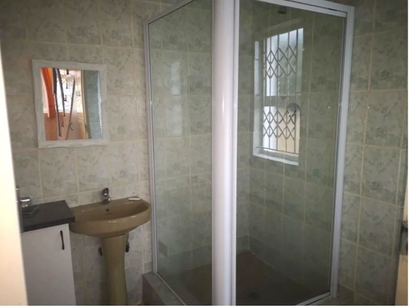 A functional bathroom featuring a corner shower enclosure, pedestal basin, and full-height wall tiling with security bars on the window.