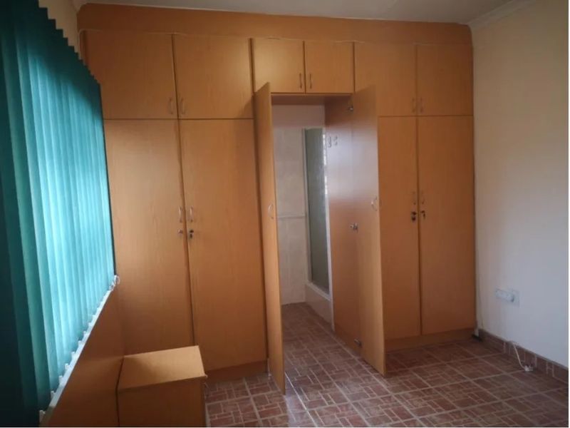A bedroom featuring extensive built-in wooden cupboards, tiled flooring, and vertical blinds, with an integrated doorway leading to an adjoining space.