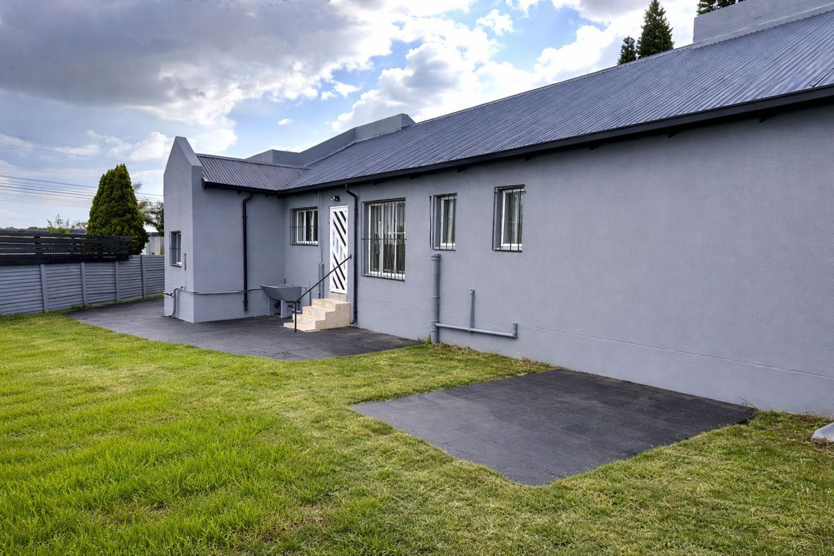 Rear view of the property featuring a grey rendered facade, corrugated metal roof, security bars on windows, and a paved patio area leading to the lawn.