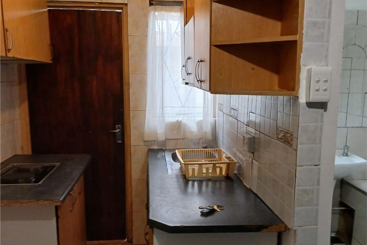 A compact, dated kitchen featuring wooden cabinetry, dark laminate countertops, and tiled walls, with an adjacent view into a small bathroom area.