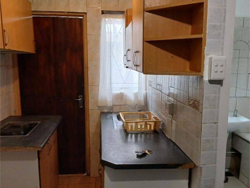 A compact, dated kitchen featuring wooden cabinetry, dark laminate countertops, and tiled walls, with an adjacent view into a small bathroom area.