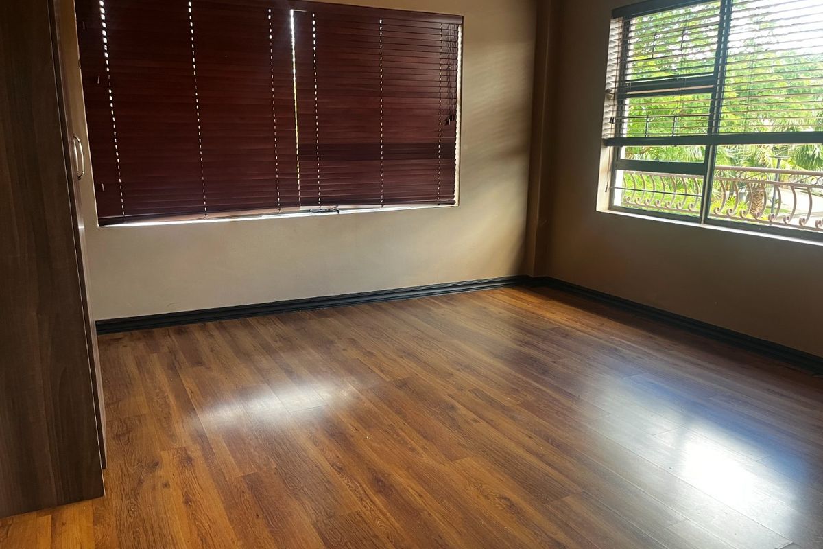 A spacious, empty bedroom featuring warm-toned laminate flooring, built-in cupboards, and large windows fitted with dark wood-look venetian blinds.