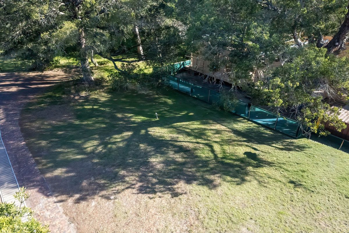 An elevated aerial view of a vacant residential plot featuring established trees, a brick-paved pathway, and perimeter fencing.