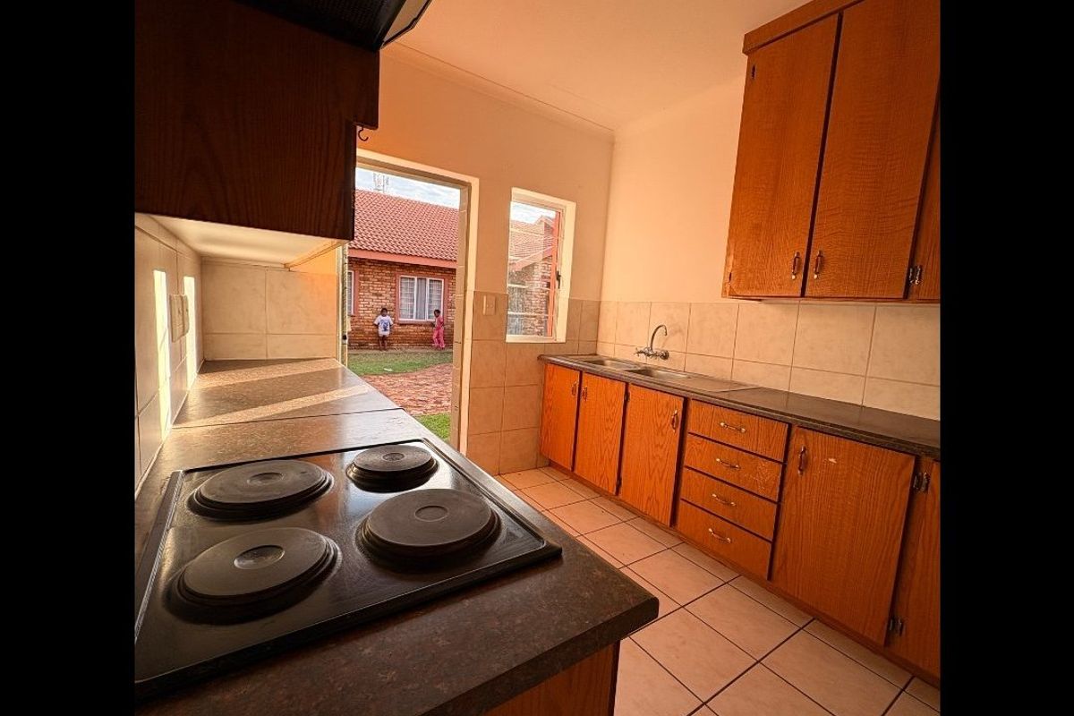 A functional kitchen featuring wooden cabinetry, an electric hob with extractor, and tiled flooring, with direct access to the rear exterior area.