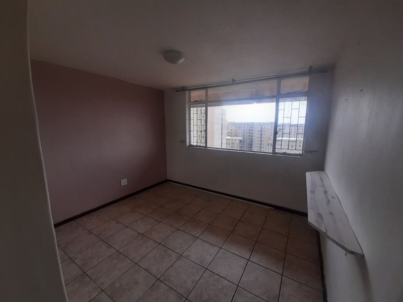 An empty bedroom featuring tiled flooring, a built-in wall shelf, and metal security bars on the window with views of the surrounding apartment buildings.