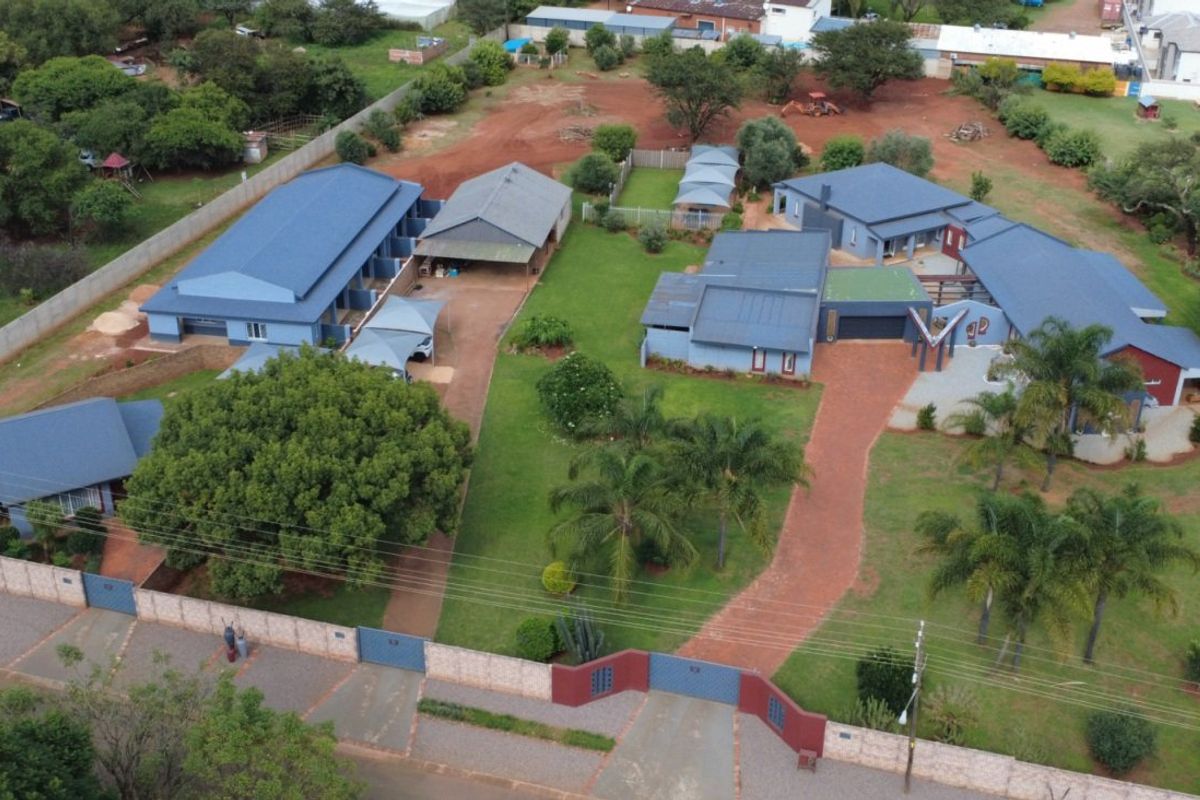 Aerial view of a spacious farm property featuring multiple buildings with metal roofs, extensive landscaped gardens, and a paved driveway with gated access.