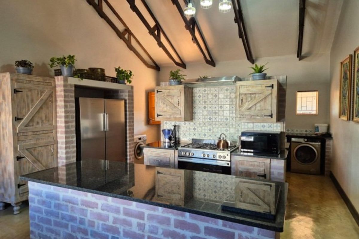 Rustic farmhouse kitchen featuring exposed timber trusses, brick island with granite countertop, gas hob, and integrated cabinetry with a tiled backsplash.