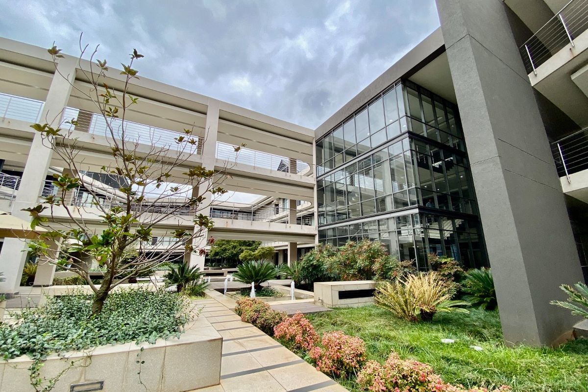 Modern commercial building exterior featuring a multi-level glass facade, landscaped garden courtyards, and elevated walkways connecting the office wings.