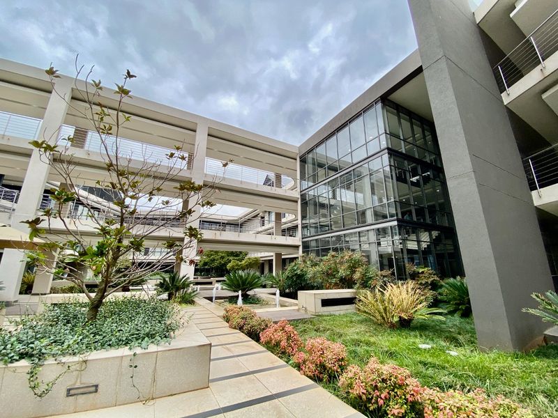 Modern commercial building exterior featuring a multi-level glass facade, landscaped garden courtyards, and elevated walkways connecting the office wings.