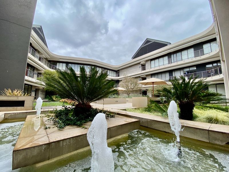 A well-maintained commercial courtyard featuring a central water fountain, stone paving, and landscaped garden beds surrounded by a multi-story office building.