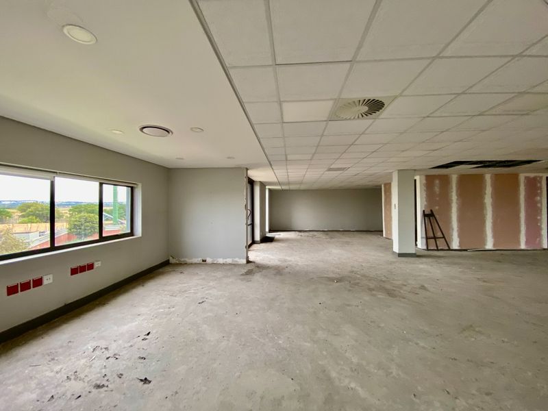 A large, vacant open-plan commercial office space featuring a suspended ceiling, recessed lighting, and unfinished wall and floor surfaces.