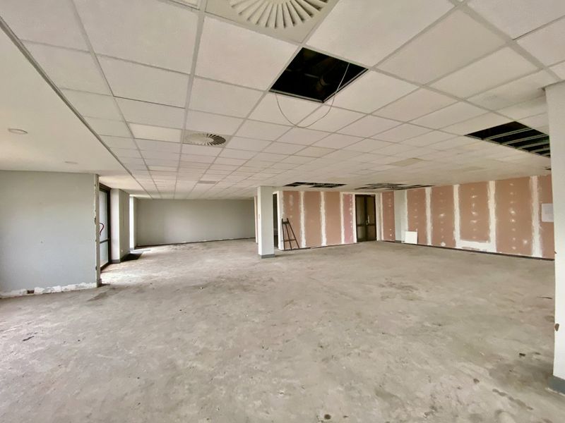 A large, open-plan commercial office space currently undergoing renovation, featuring a suspended ceiling with missing tiles and unfinished wall surfaces.