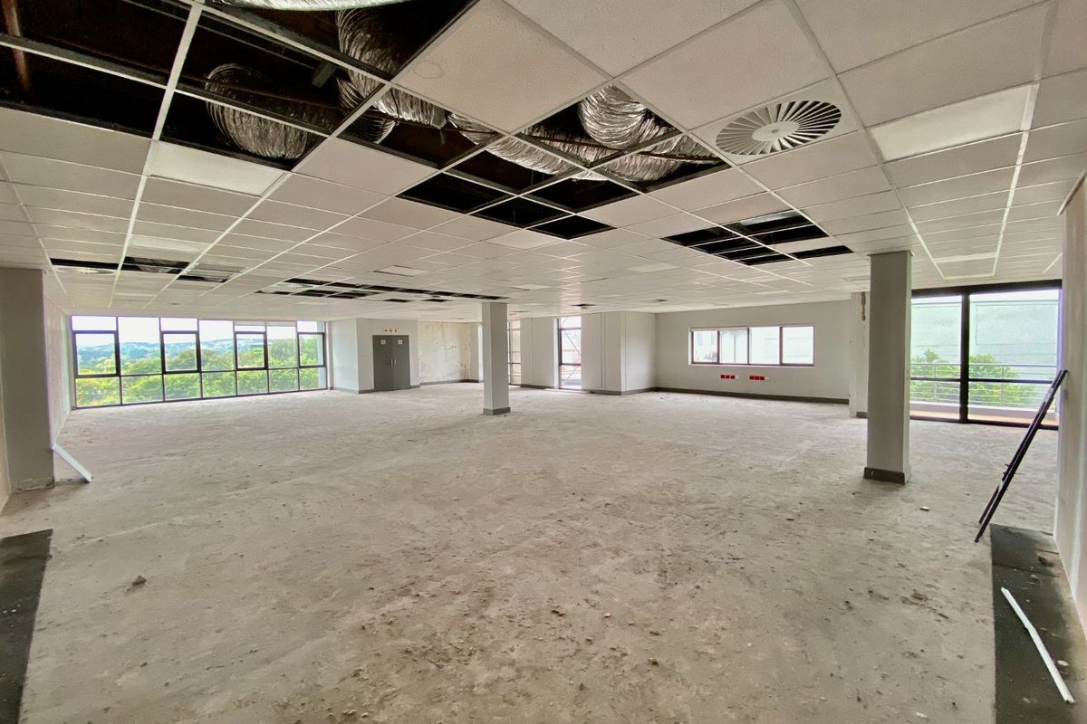 A large, open-plan commercial office space featuring expansive windows, structural columns, and an unfinished ceiling with exposed HVAC ducting.