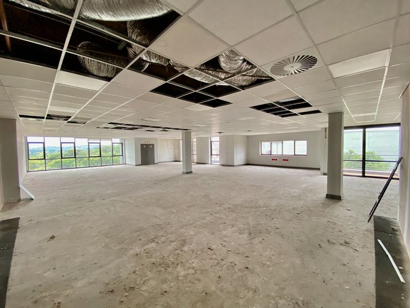 A large, open-plan commercial office space featuring expansive windows, structural columns, and an unfinished ceiling with exposed HVAC ducting.