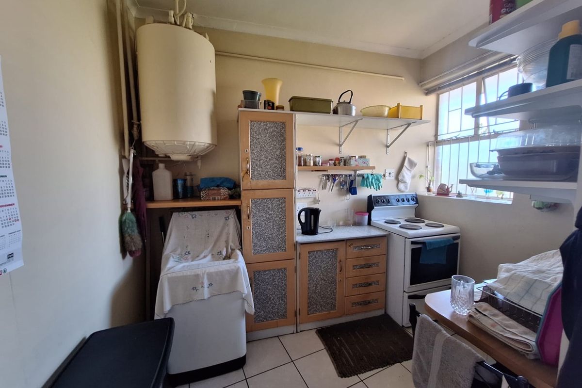 A compact kitchen featuring dated cabinetry, an electric stove, and an exposed water heater, with tiled flooring and security bars on the window.