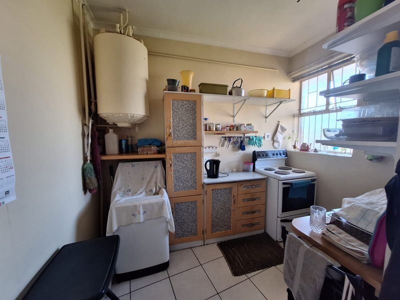 A compact kitchen featuring dated cabinetry, an electric stove, and an exposed water heater, with tiled flooring and security bars on the window.