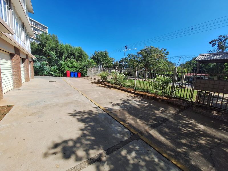 A paved rear exterior area featuring a concrete driveway, metal security fencing with razor wire, and access to the building's garage section.