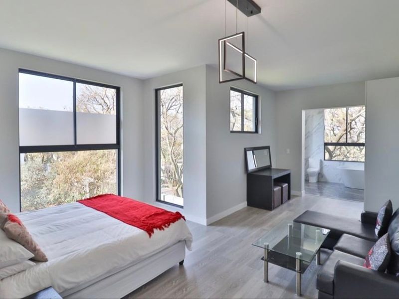 Spacious modern bedroom featuring laminate flooring, large black-framed windows with natural light, and an open-plan en-suite bathroom.
