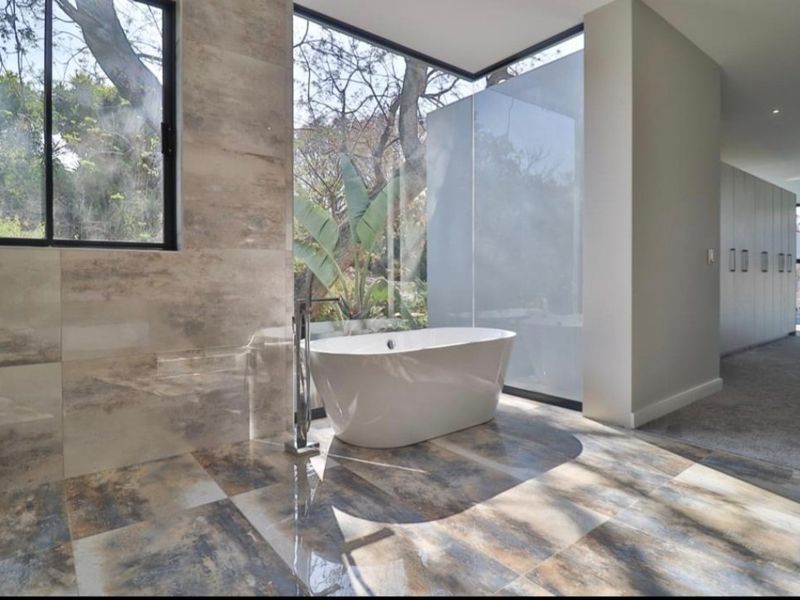 Modern bathroom featuring a sleek freestanding bathtub, large floor-to-ceiling windows with garden views, and high-quality tiled finishes.