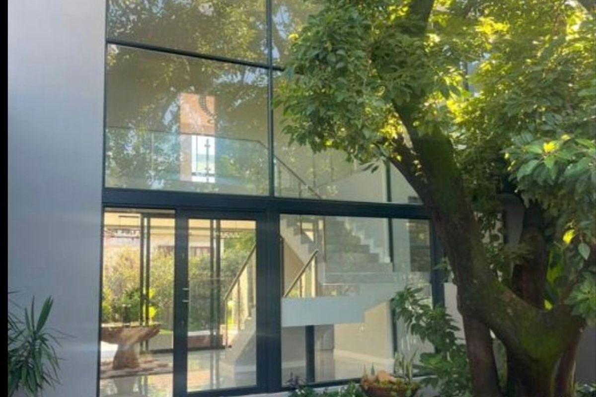 Modern rear elevation featuring a striking double-volume glass facade, paved courtyard, and a contemporary staircase visible through the expansive windows.