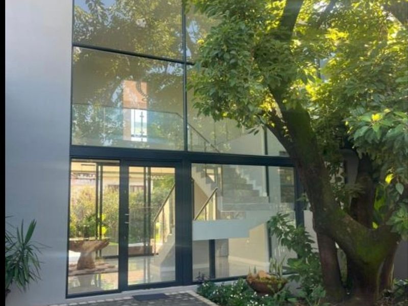Modern rear elevation featuring a striking double-volume glass facade, paved courtyard, and a contemporary staircase visible through the expansive windows.