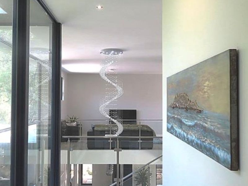 A modern, light-filled hallway featuring a glass balustrade, contemporary chandelier, and large windows overlooking the lower level.