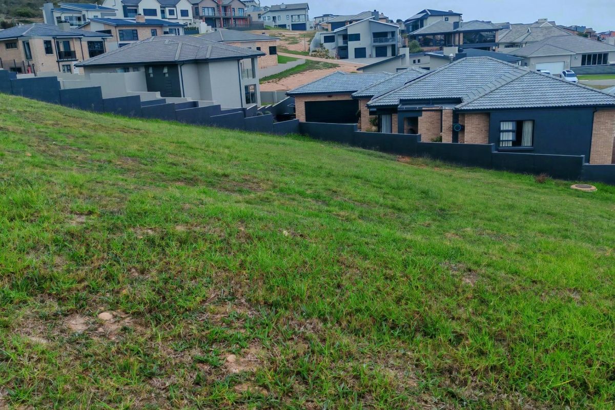 A sloped, grass-covered vacant residential plot situated within an established neighborhood, bordered by a modern boundary wall and adjacent to existing residential properties.