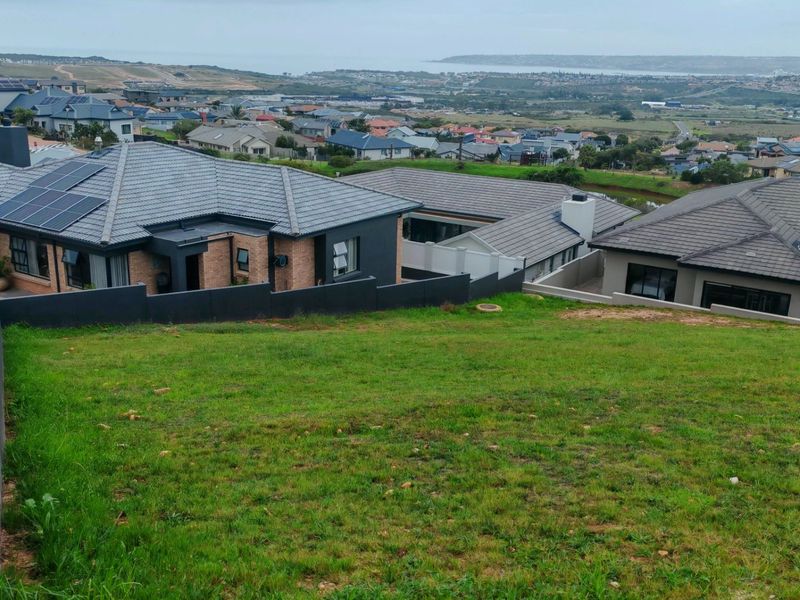A view of a sloped residential plot overlooking a suburban neighborhood and distant coastal landscape.