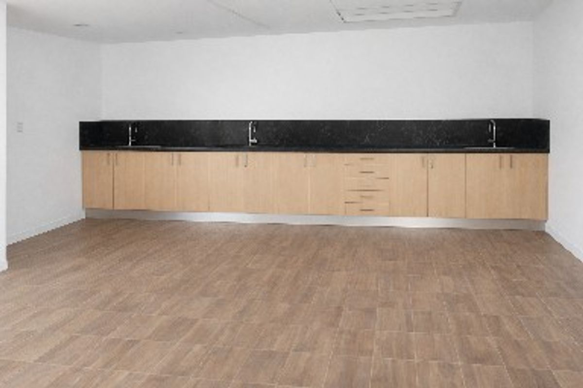 A clean, empty commercial kitchen space featuring extensive light wood cabinetry, dark stone countertops, multiple sinks, and durable tiled flooring.