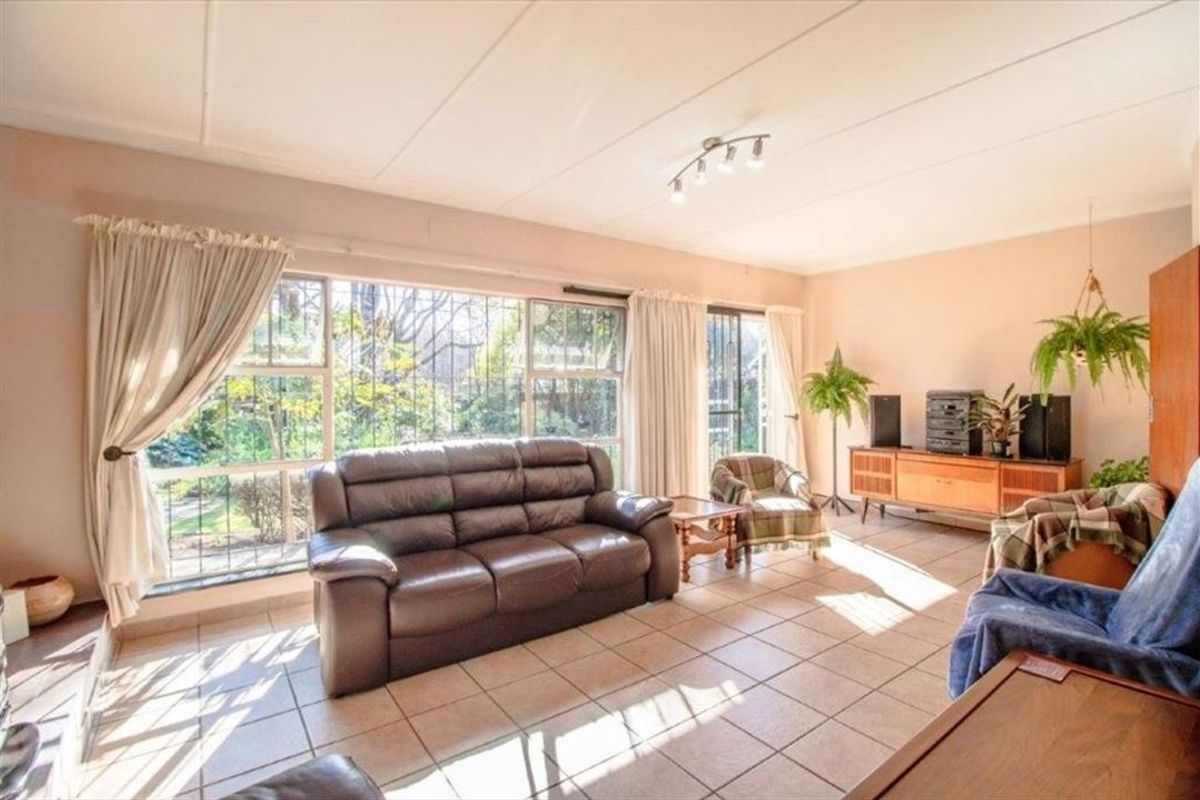 A spacious living room featuring large windows with security bars, tiled flooring, and neutral wall finishes, offering plenty of natural light.