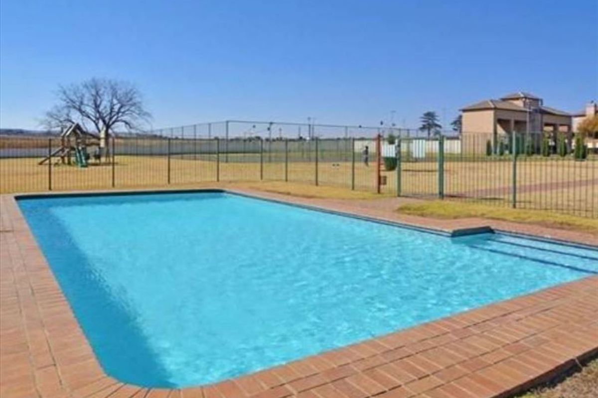 A communal outdoor swimming pool area featuring brick coping, surrounded by metal fencing with a playground visible in the background.