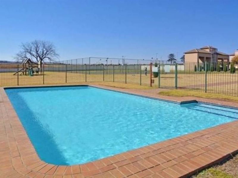 A communal outdoor swimming pool area featuring brick coping, surrounded by metal fencing with a playground visible in the background.
