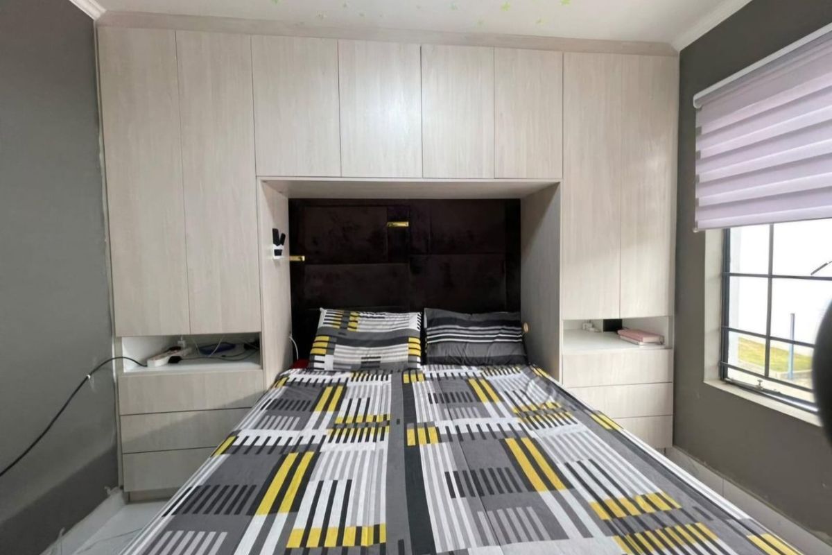 A bedroom featuring a large, light-wood built-in wardrobe unit with integrated headboard and storage, complemented by a window with a roller blind.