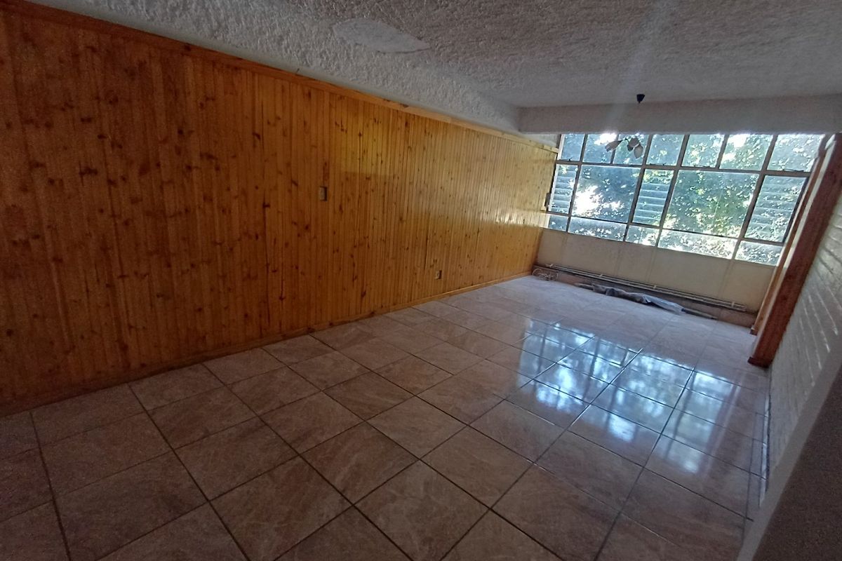 An empty living room featuring dated wood wall paneling, tiled flooring, and large windows with louvre panes, offering a blank canvas for renovation.