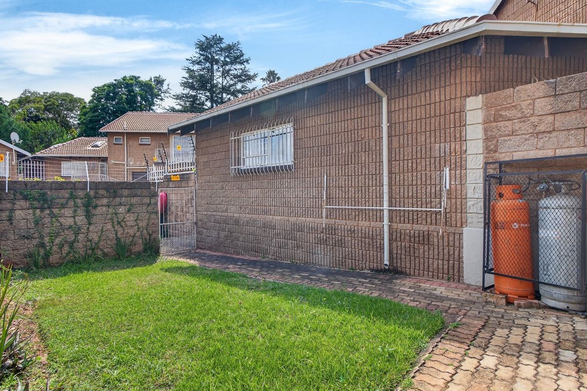 Rear exterior view of the townhouse featuring a brick facade, paved walkway, small lawn area, and a secure gas cylinder storage cage.