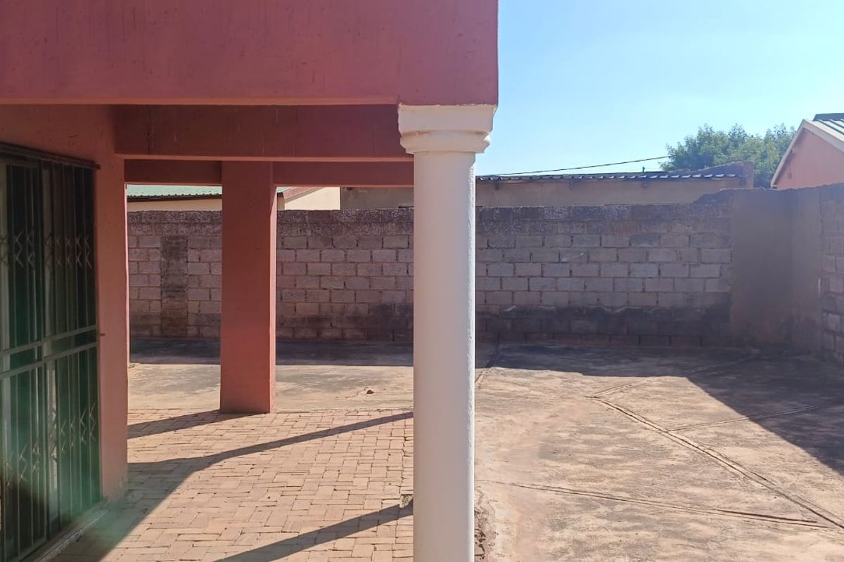 A covered outdoor patio area featuring brick paving, structural columns, and a security gate, leading to an enclosed rear yard with boundary walls.