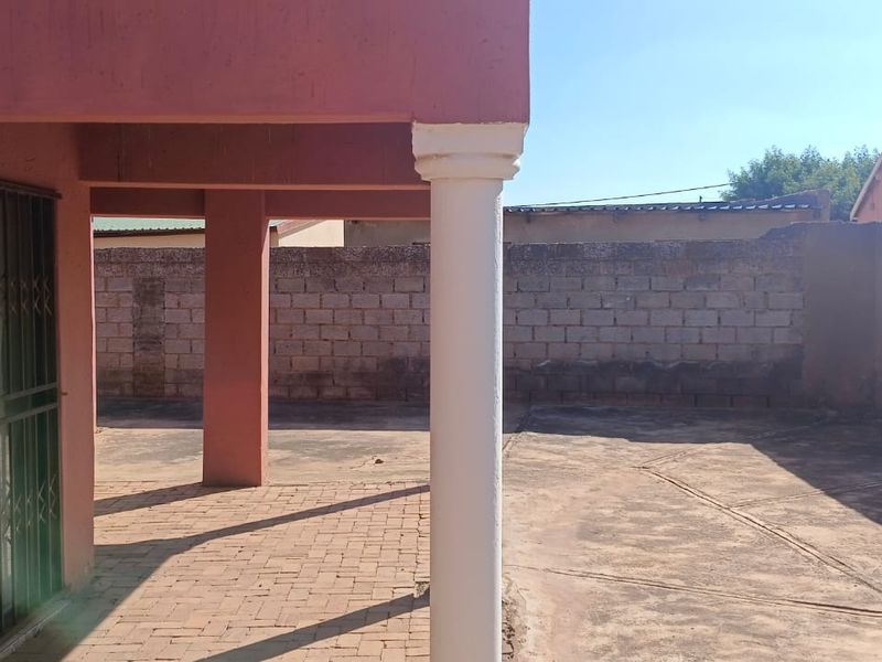 A covered outdoor patio area featuring brick paving, structural columns, and a security gate, leading to an enclosed rear yard with boundary walls.