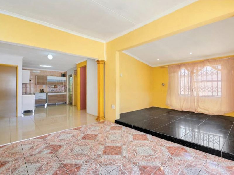 Open-plan living area featuring a raised platform, decorative columns, and an adjoining kitchen space with built-in cabinetry and tiled flooring throughout.
