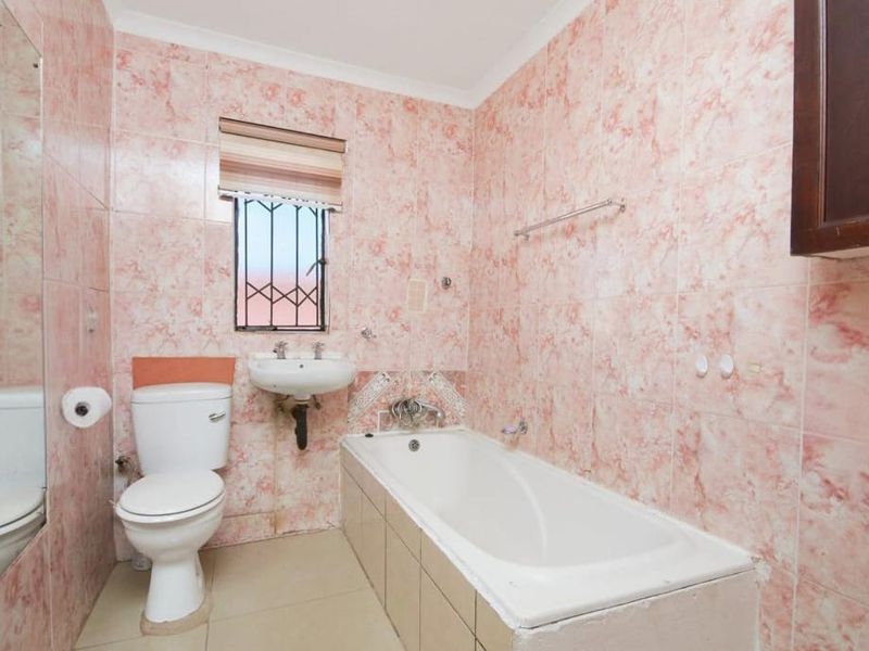 A dated bathroom featuring pink marble-effect wall tiles, a standard bathtub, pedestal sink, and toilet, with security bars on the window.