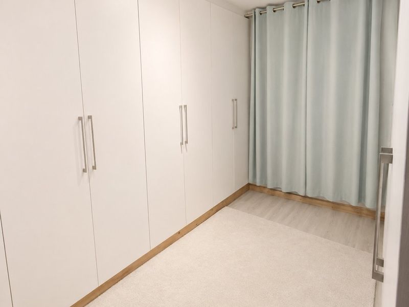 A clean, modern bedroom featuring extensive built-in white wardrobes, light-toned laminate flooring, and contemporary track lighting.