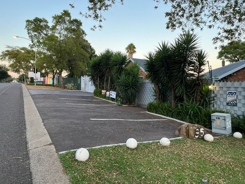 Street view showing an asphalt parking area, concrete boundary wall with electric fencing, and established landscaping along the property frontage.