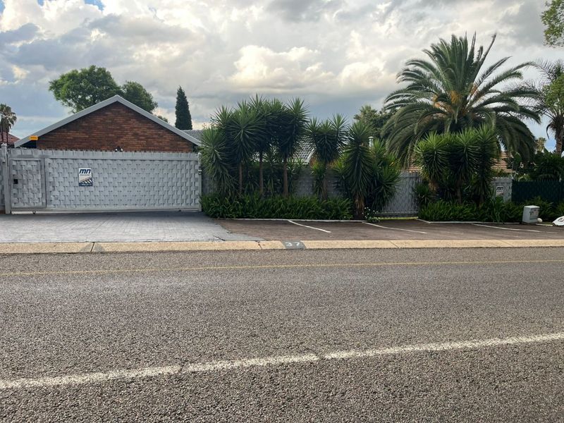 Street view of the property featuring a brick facade, secure gated entrance, paved driveway, and established garden landscaping.
