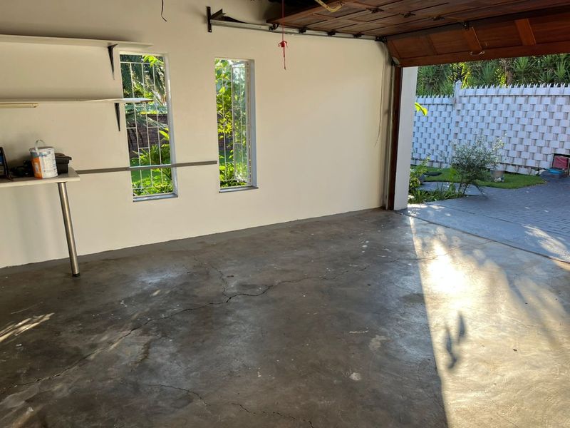 A spacious, empty garage featuring concrete flooring with visible cracks, security-barred windows, and built-in shelving, offering direct access to the rear garden area.