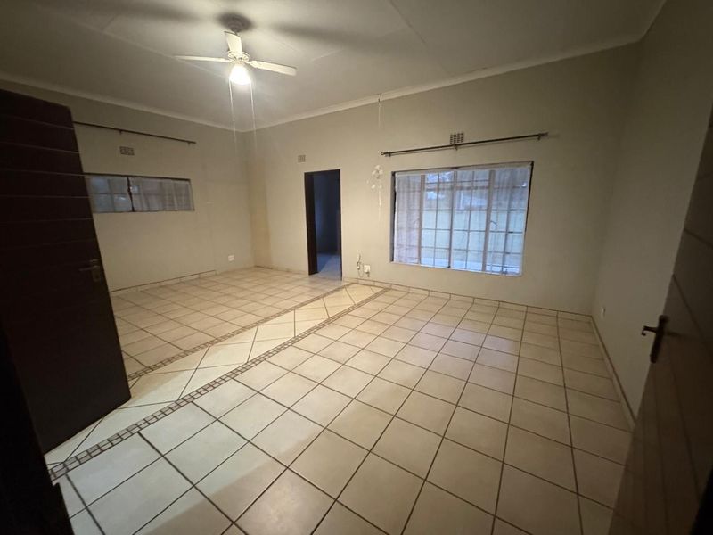 A spacious, empty living room featuring neutral tiled flooring, a ceiling fan, and large windows fitted with security bars.