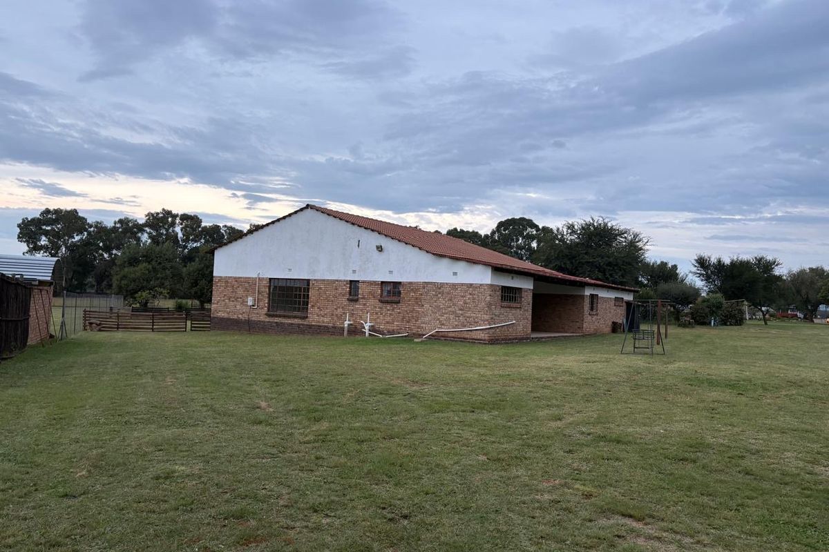 Rear view of a single-story brick house featuring a tiled roof, steel-framed windows, and a covered patio area set on a large grass lawn.