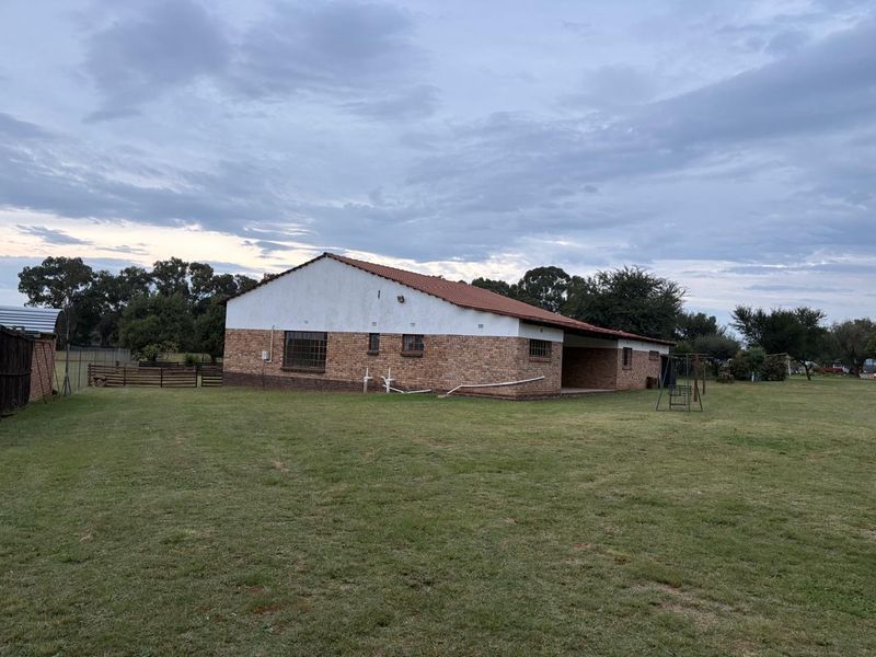 Rear view of a single-story brick house featuring a tiled roof, steel-framed windows, and a covered patio area set on a large grass lawn.