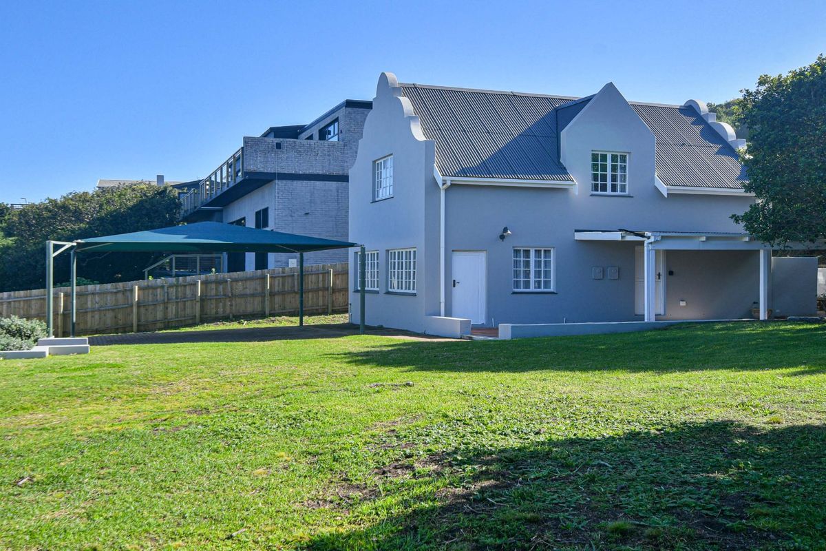 Rear view of a Cape Dutch style house featuring a gabled roof, covered patio area, and a spacious lawn with a shade sail structure.