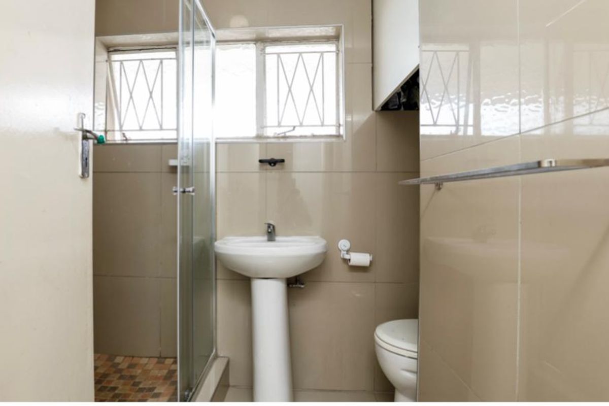 A functional bathroom featuring a pedestal basin, toilet, and a glass-enclosed shower with tiled walls and security bars on the window.