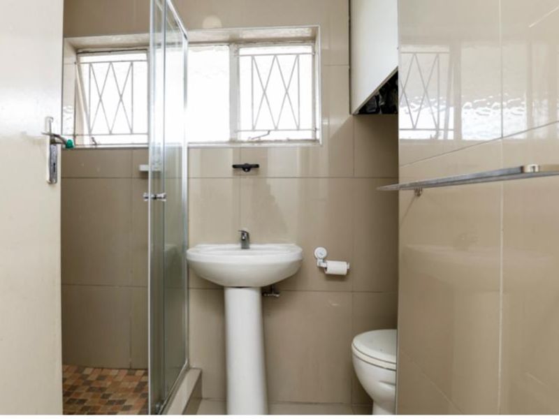 A functional bathroom featuring a pedestal basin, toilet, and a glass-enclosed shower with tiled walls and security bars on the window.