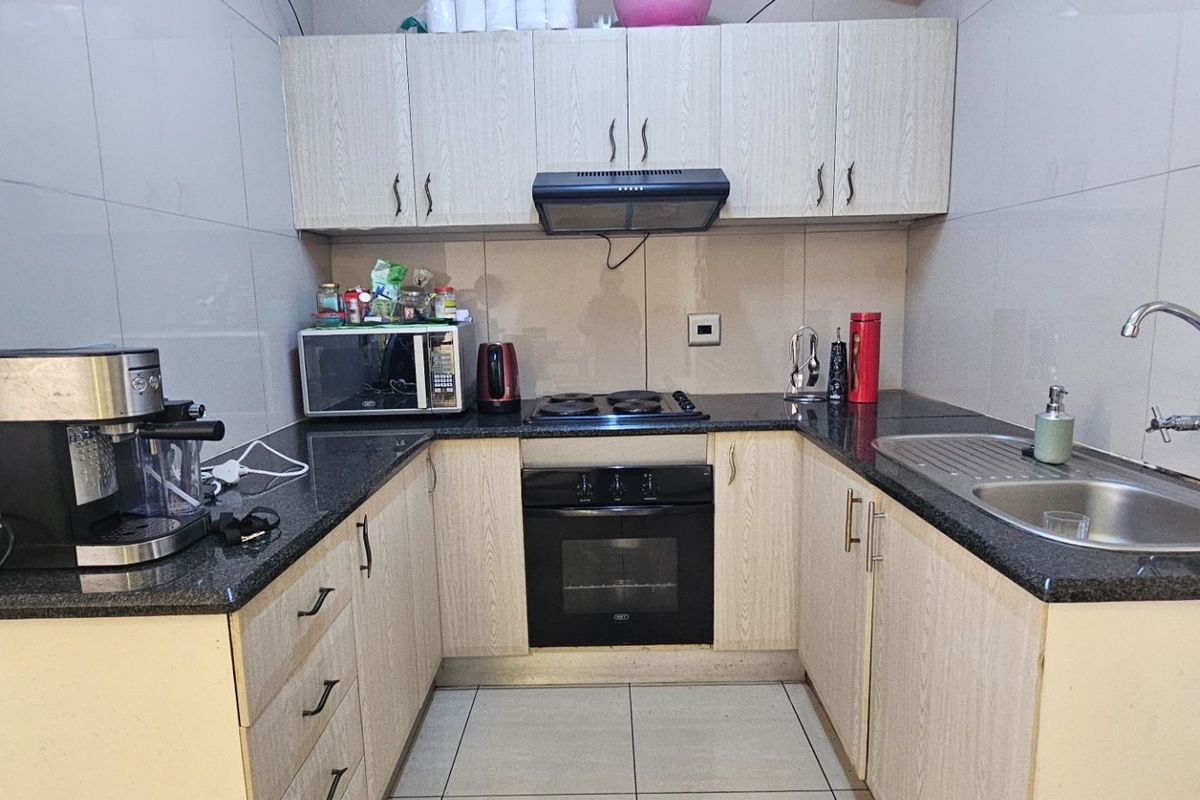 Compact kitchen featuring light wood-finish cabinetry, granite countertops, built-in oven, electric hob, extractor fan, and a stainless steel sink with tiled finishes.
